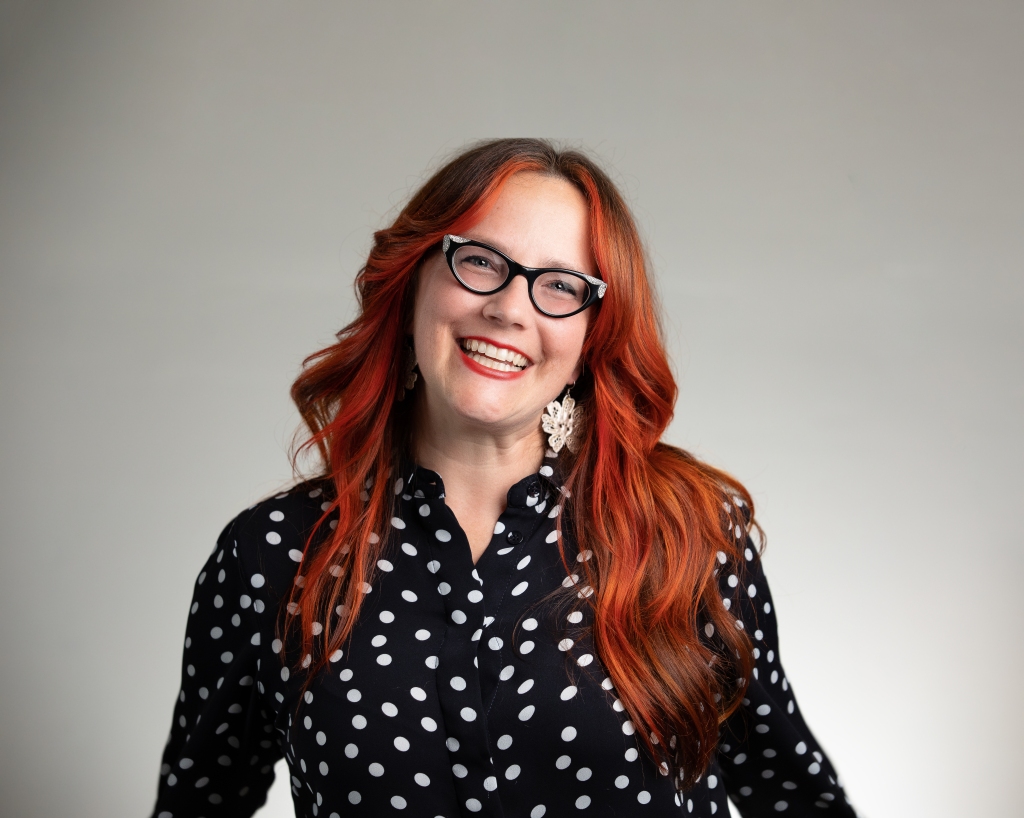 Meet Amy Higgins. A creative gal who loves funky glasses like the vintage cat eyes she's wearing in this photo, wearing polka dots, like the black shirt with white dots she's wearing here, and expressing herself with fun hairstyles, like her bright red hair here in this photo.